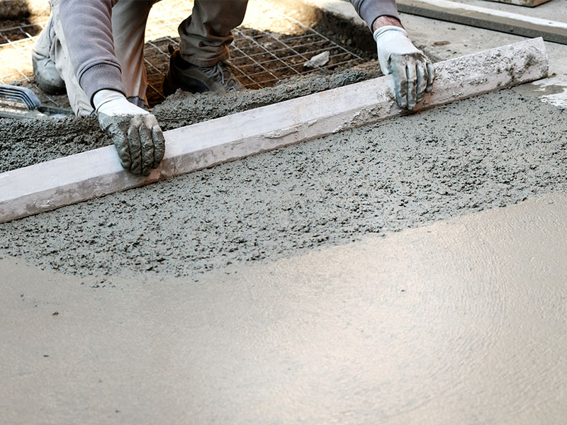 Das Bild zeigt die gezielte Arbeit eines Handwerkers beim Abziehen von frisch aufgetragenem Estrich. Mit einer Abziehlatte wird die Oberfläche gleichmäßig geglättet, um eine perfekte Basis für den Bodenbelag zu schaffen. Im Hintergrund ist die Baustelle mit sichtbarer Armierung zu erkennen, die zur Stabilität des Estrichs beiträgt. Die Handschuhe des Handwerkers verdeutlichen die notwendige Sorgfalt und den direkten Kontakt mit dem Material.