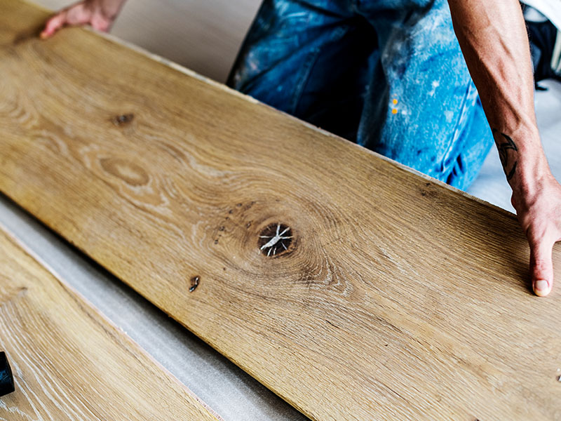 Das Bild zeigt die detailreiche Struktur einer hochwertigen Landhausdiele aus Holz. Die markante Maserung und natürliche Astlöcher betonen den rustikalen Charakter des Materials. Ein Handwerker legt die Diele mit Präzision und Sorgfalt, was auf die hohe Qualität und den individuellen Charme von Parkettböden hinweist.