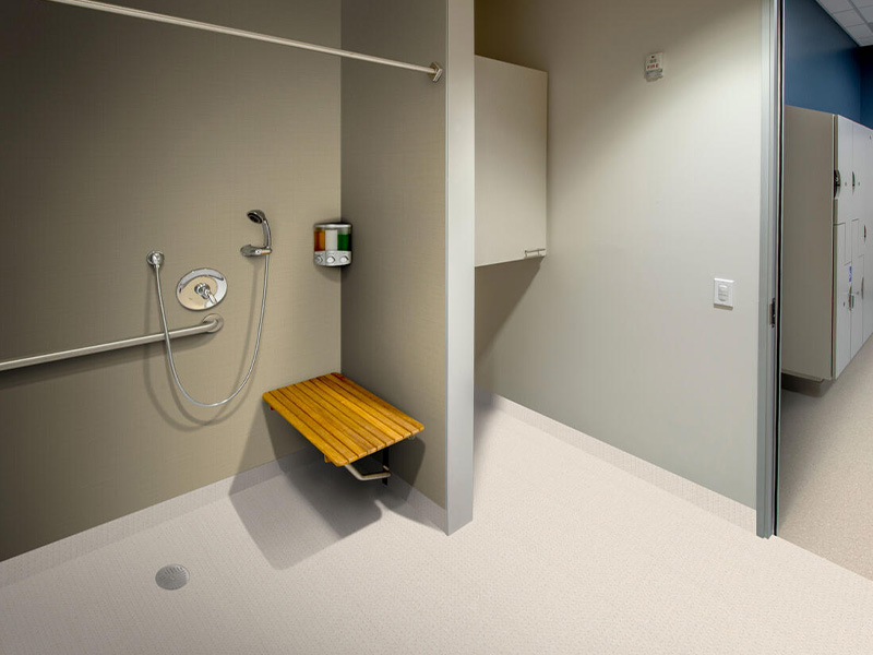 Das Bild zeigt eine barrierefreie Duschkabine mit klappbarem Holzsitz, Handbrause und Haltegriffen für eine sichere Nutzung. Der Boden ist mit einer rutschfesten Wannenformverlegung ausgestattet, die nahtlos in den Wandbereich übergeht, um Hygiene und Sicherheit zu gewährleisten. Der angrenzende Bereich führt in eine Umkleidekabine mit Schließfächern und klar strukturierter Einrichtung, ideal für öffentliche oder private Pflegeeinrichtungen.