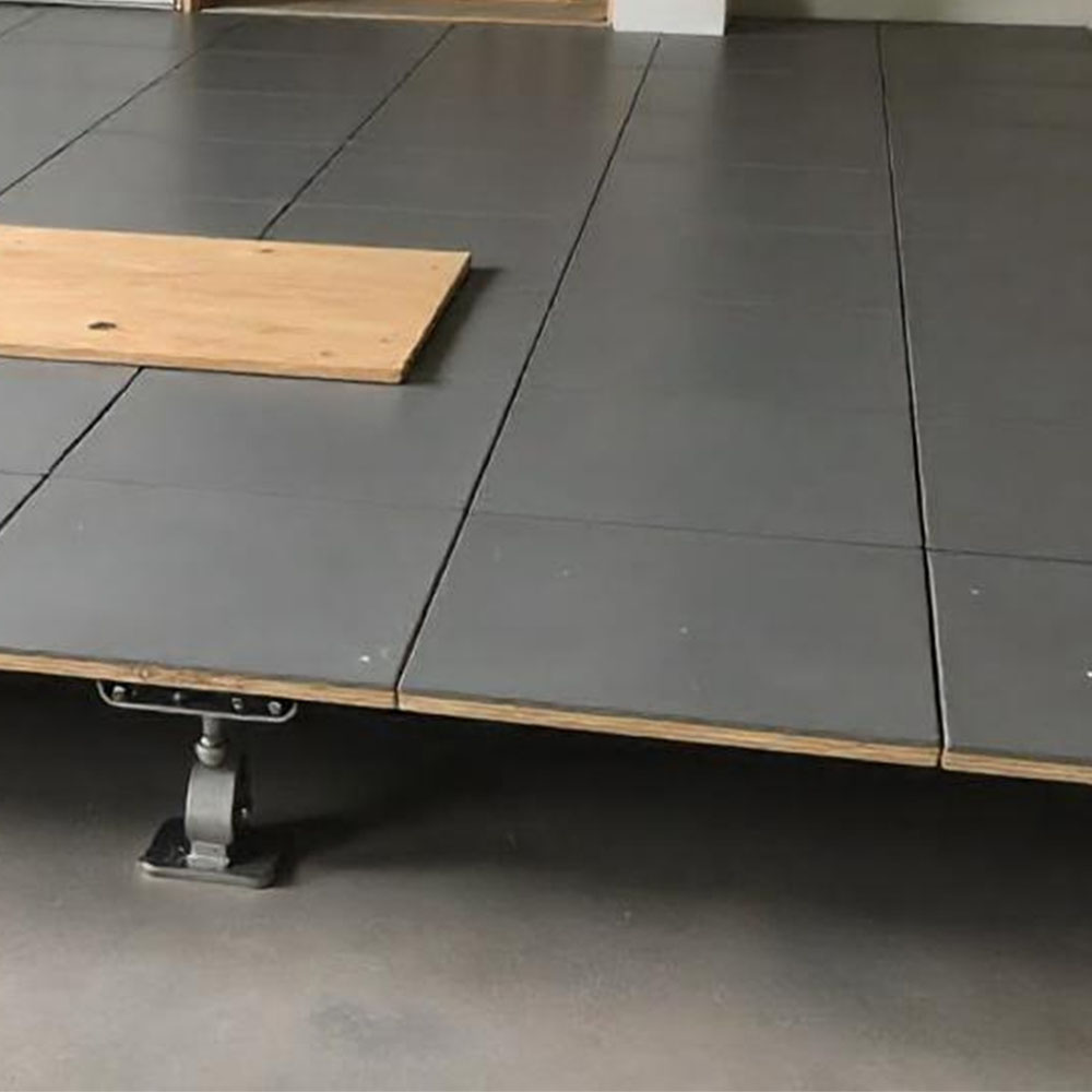 Das Bild zeigt den Aufbau eines Hohlraumbodensystems. Die Bodenplatten sind präzise auf höhenverstellbaren Stelzen montiert, die eine stabile Basis und gleichzeitig Platz für Kabel, Rohre und andere Installationen darunter schaffen. Eine lose Holzplatte verdeutlicht die Flexibilität der Konstruktion und den modularen Charakter des Systems. Ideal für moderne Büro- oder Technikräume, in denen eine verdeckte Verkabelung erforderlich ist.